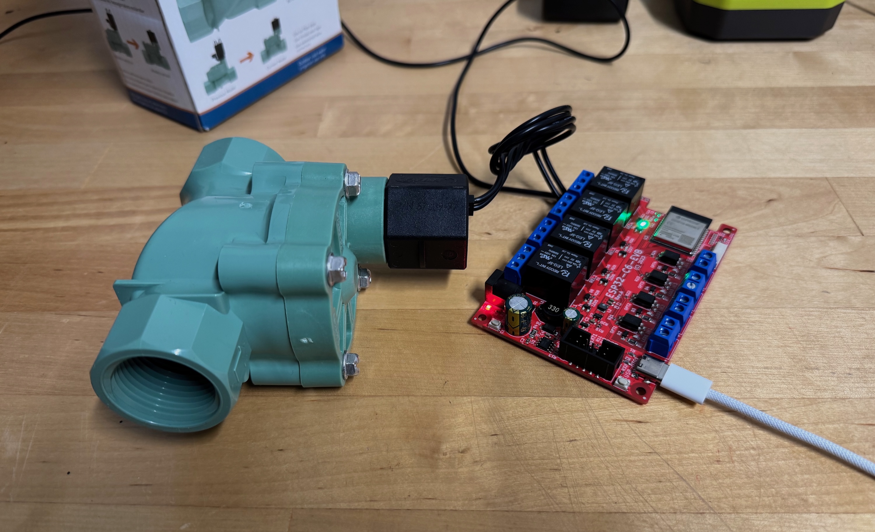 An Olimex ESP32-C6-EVB relay board on a workbench, powered over USB-C with one relay's status LED lit, its screw terminal wired to the coil of a green brass sprinkler solenoid valve sitting beside it.
